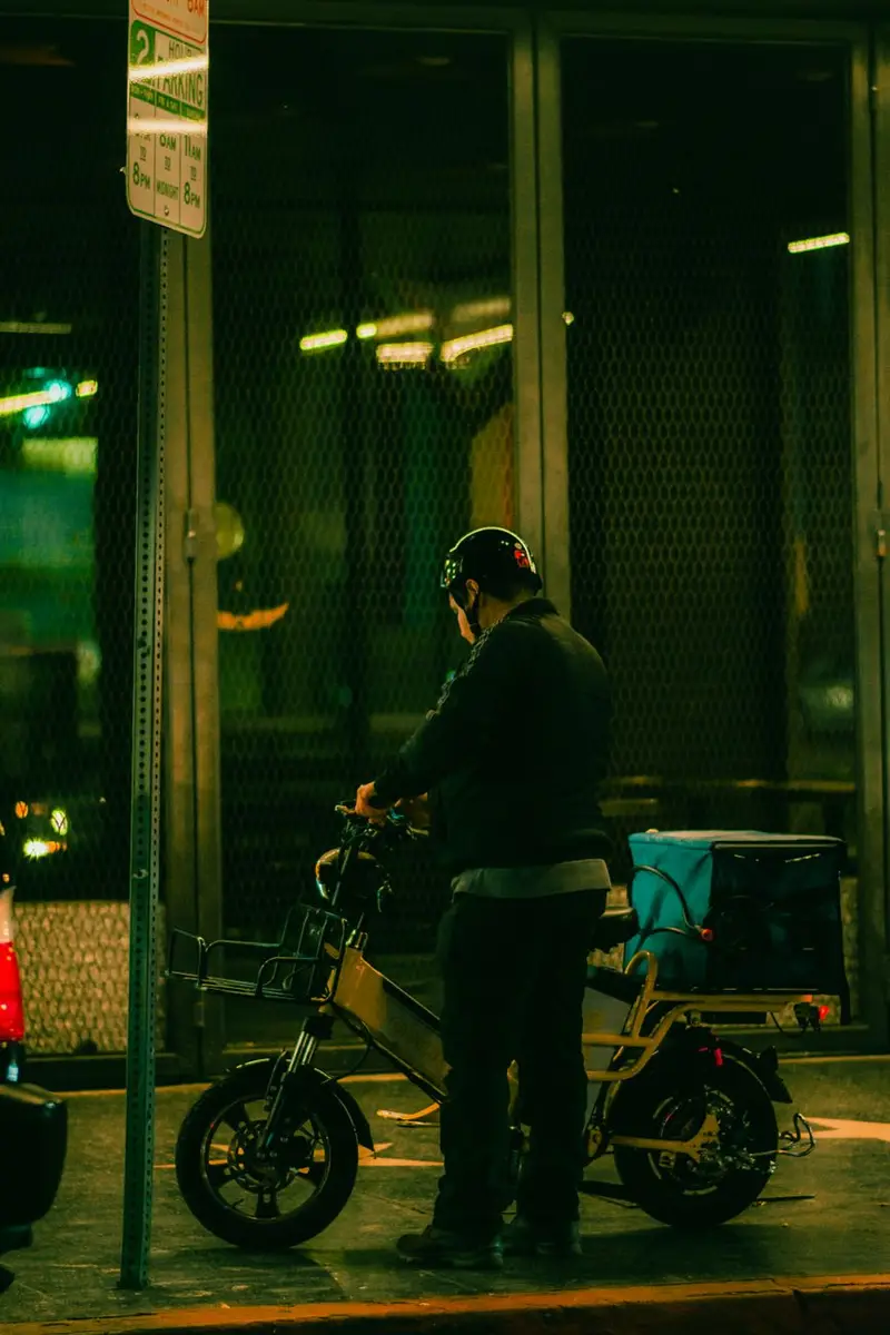 Delivery rider on an electric bike with a cargo rack, stopped outside a row of shops